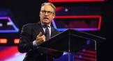 Eric Metaxas speaks, wearing glasses and suit, on a conference stage with neon blue and red background