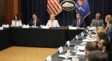 Officials sit in tables in an office with a Justice Department emblem and US flag.
