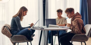 Three teenagers using laptops and smartphones while doing homework.
