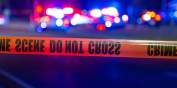 Police lights flash behind tape at a crime scene at night.