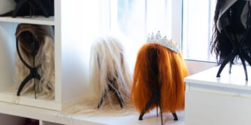 Wigs of various colors and styles, including a tiara, sit on a white shelf in a bright dressing room, likely belonging to a performer or entertainer.