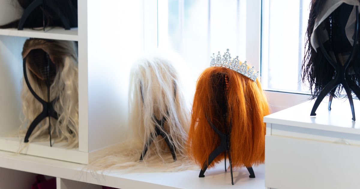 Wigs of various colors and styles, including a tiara, sit on a white shelf in a bright dressing room, likely belonging to a performer or entertainer.