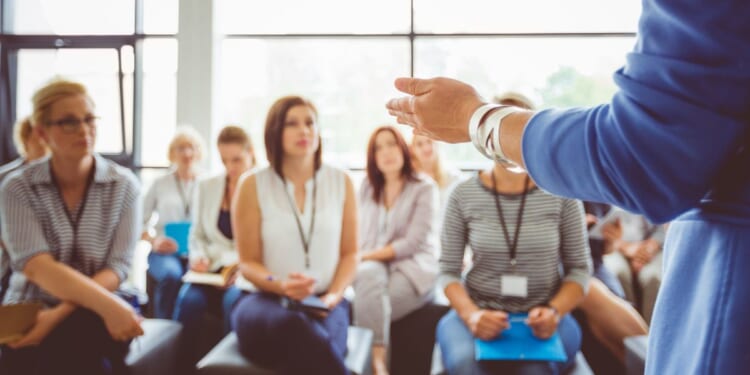 The hand of a trainer addressing a group of females sitting in a conference hall.