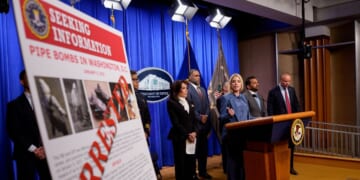 A poster reads "ARRESTED" in bold letters as Attorney General Pam Bondi speaks during a news conference on an arrest of a suspect in the January 6th pipe bomb case at the Department of Justice on Dec. 4, 2025, in Washington, D.C.