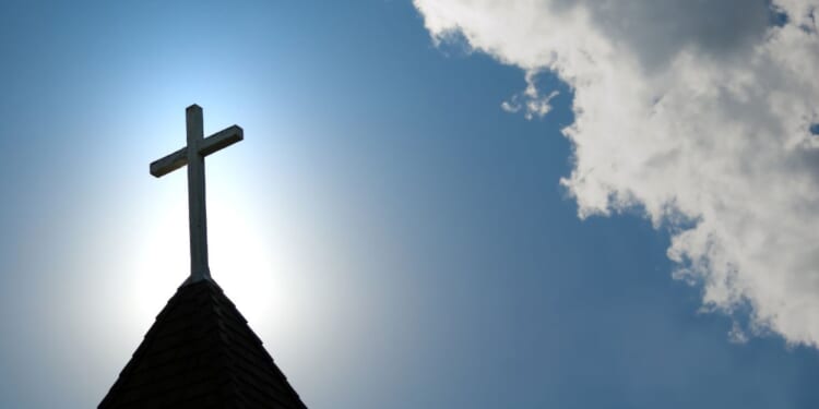A wood cross sits on top of an old church steeple in front of a blue sky.