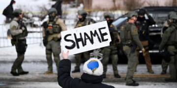 An onlooker holds a sign that reads "Shame" as members of law enforcement work the scene following a suspected shooting by an ICE agent during federal law enforcement operations on Jan. 7, 2026, in Minneapolis, Minnesota.
