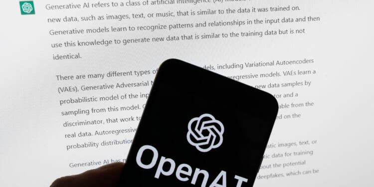 The OpenAI logo is displayed on a mobile phone in front of a computer screen with output from ChatGPT on March 21, 2023, in Boston, Massachusetts.