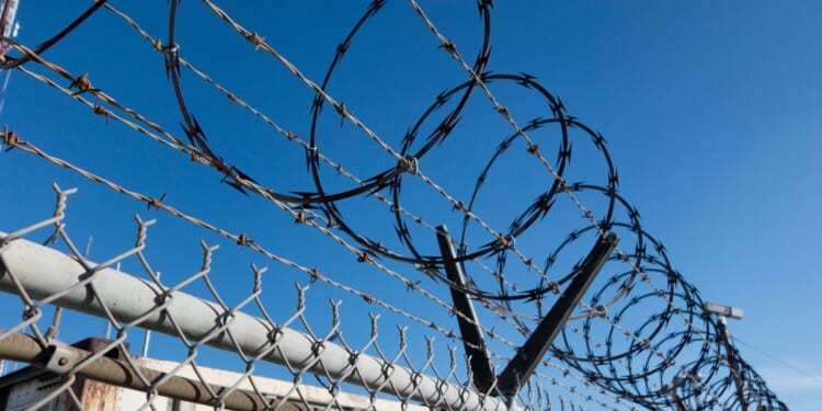 Barbed and razor wire is seen on a fence with a security camera in the background.