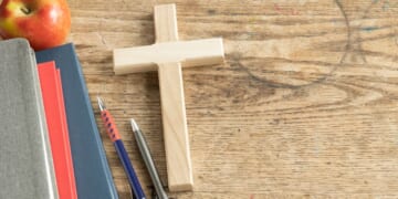 A wooden cross sits on a vintage wooden desk next to some pencils and a stack of notebooks.