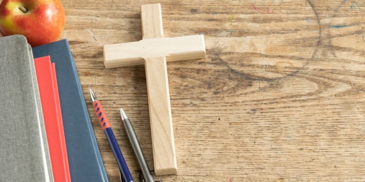 A wooden cross sits on a vintage wooden desk next to some pencils and a stack of notebooks.