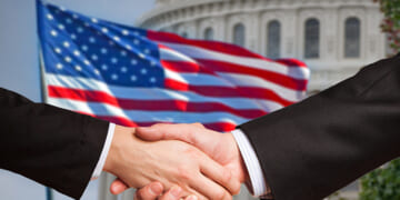 Two men shake hands as the American flag waves behind the United States Capitol rotunda.