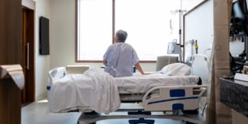 A photo illustration depicts an older woman seated on a hospital bed alone in her room.