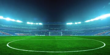 An illuminated soccer stadium at night with a green field and empty stands, ready for an exciting match under bright blue floodlights.